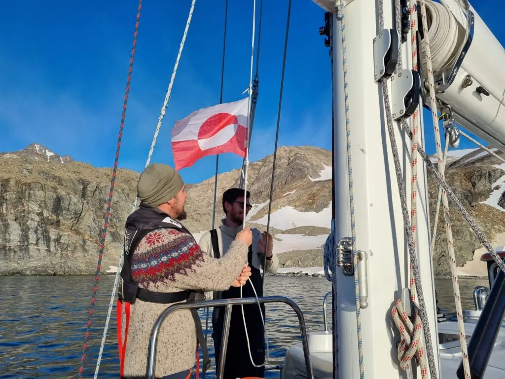 Grønlands flagg symboliserer en oppstående sol over isen, der det hvite står for lyset og det røde for varmens tilstedeværelse ved midtsommer. Her heises gjesteflagget i første naturhavn i Neil Bugt.